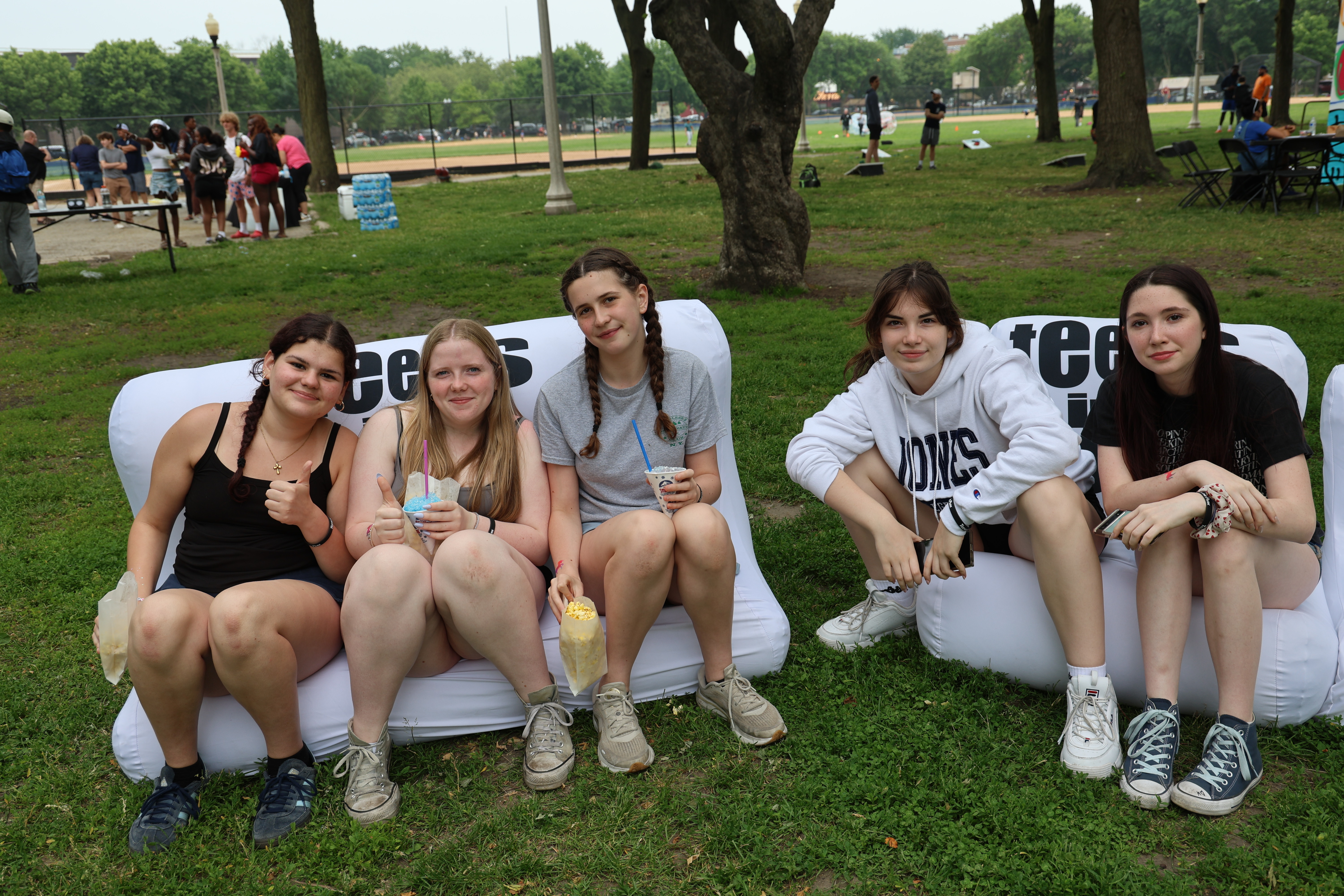 Four teenage girls sit on inflatable chairs on grass, enjoying snacks and drinks.